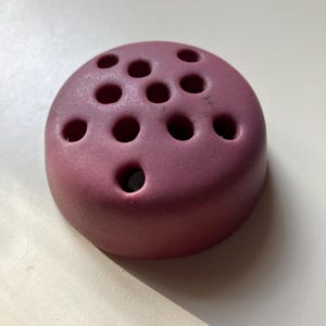May include: A round, plum-colored flower frog with multiple holes for arranging stems. The ceramic floral arranger is designed to hold flowers in place, creating beautiful bouquets. The holes are of varying sizes.
