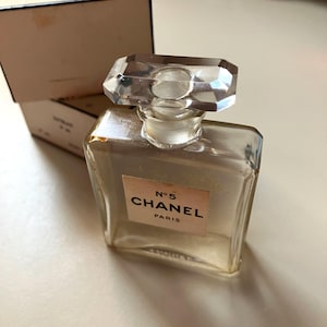 May include: A clear glass perfume bottle with a faceted crystal stopper. The bottle has a rectangular shape with a label that reads "N&deg;5 CHANEL PARIS". The box behind the bottle has the word "CHANEL" printed on it.