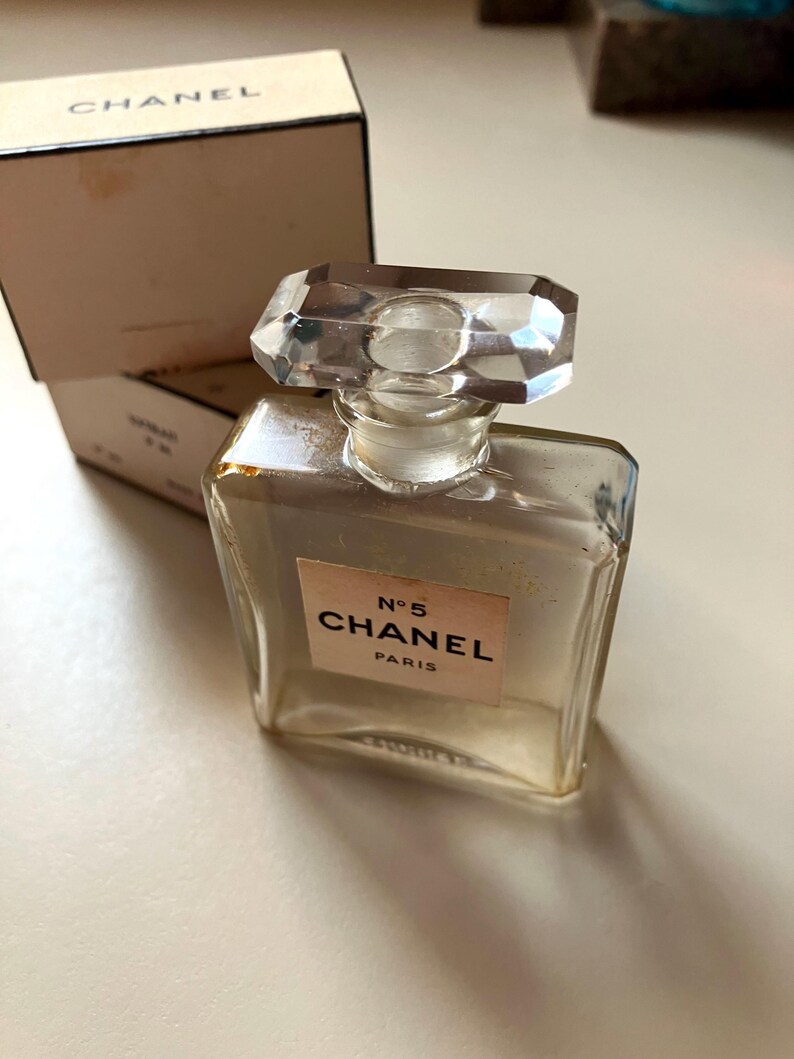 May include: A clear glass perfume bottle with a faceted crystal stopper. The bottle has a rectangular shape with a label that reads "N&deg;5 CHANEL PARIS". The box behind the bottle has the word "CHANEL" printed on it.