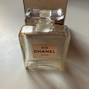 May include: A clear, rectangular Chanel No. 5 perfume bottle with a faceted, rectangular stopper. The label reads "N&deg;5 CHANEL PARIS" on a light peach background. A matching box is in the background.