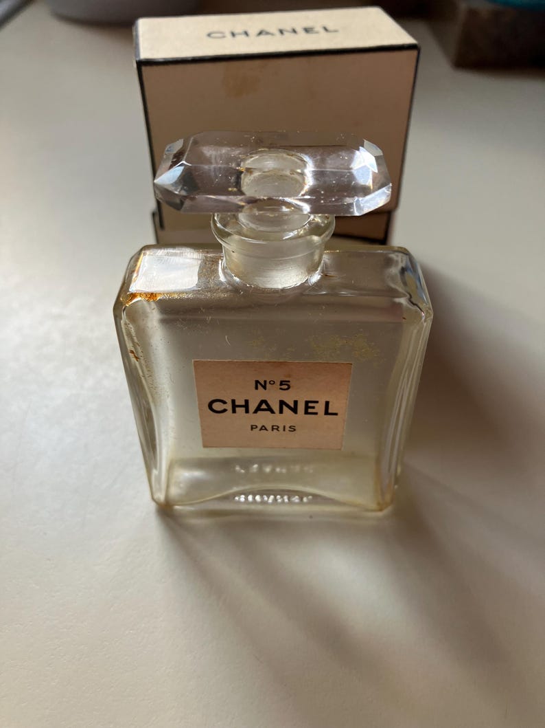 May include: A clear, rectangular Chanel No. 5 perfume bottle with a faceted, rectangular stopper. The label reads "N&deg;5 CHANEL PARIS" on a light peach background. A matching box is in the background.