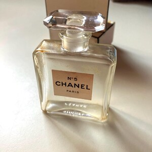 May include: A clear glass perfume bottle with a rectangular shape and a faceted, clear stopper. The label reads "N&deg;5 CHANEL PARIS" in black text on a beige background. The bottle is in front of a beige box.