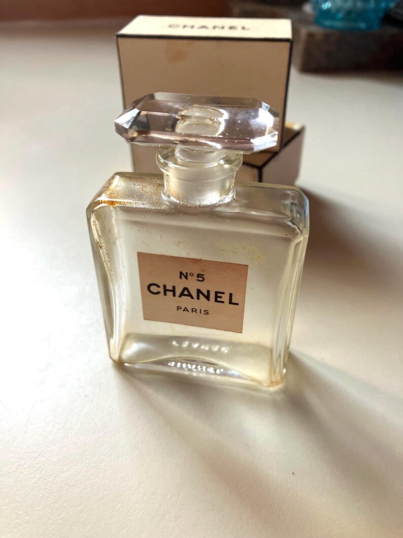 May include: A clear glass perfume bottle with a rectangular shape and a faceted, clear stopper. The label reads "N&deg;5 CHANEL PARIS" in black text on a beige background. The bottle is in front of a beige box.