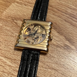 May include: A gold-toned rectangular wristwatch with a skeleton dial, showcasing the intricate inner workings. The watch features a black leather band. The watch is displayed on a wooden surface.