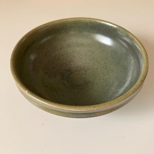 May include: A round ceramic bowl with a smooth, glossy finish in shades of olive green and muted sage. The interior features a subtle mottled texture with darker green speckles, while the rim is slightly raised and defined by a lighter tone.