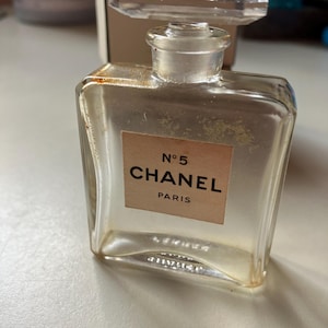May include: A clear glass perfume bottle with a rectangular shape and a clear, faceted stopper. The label reads "N&deg;5 CHANEL PARIS" in black text on a beige background. The bottle appears to be vintage.