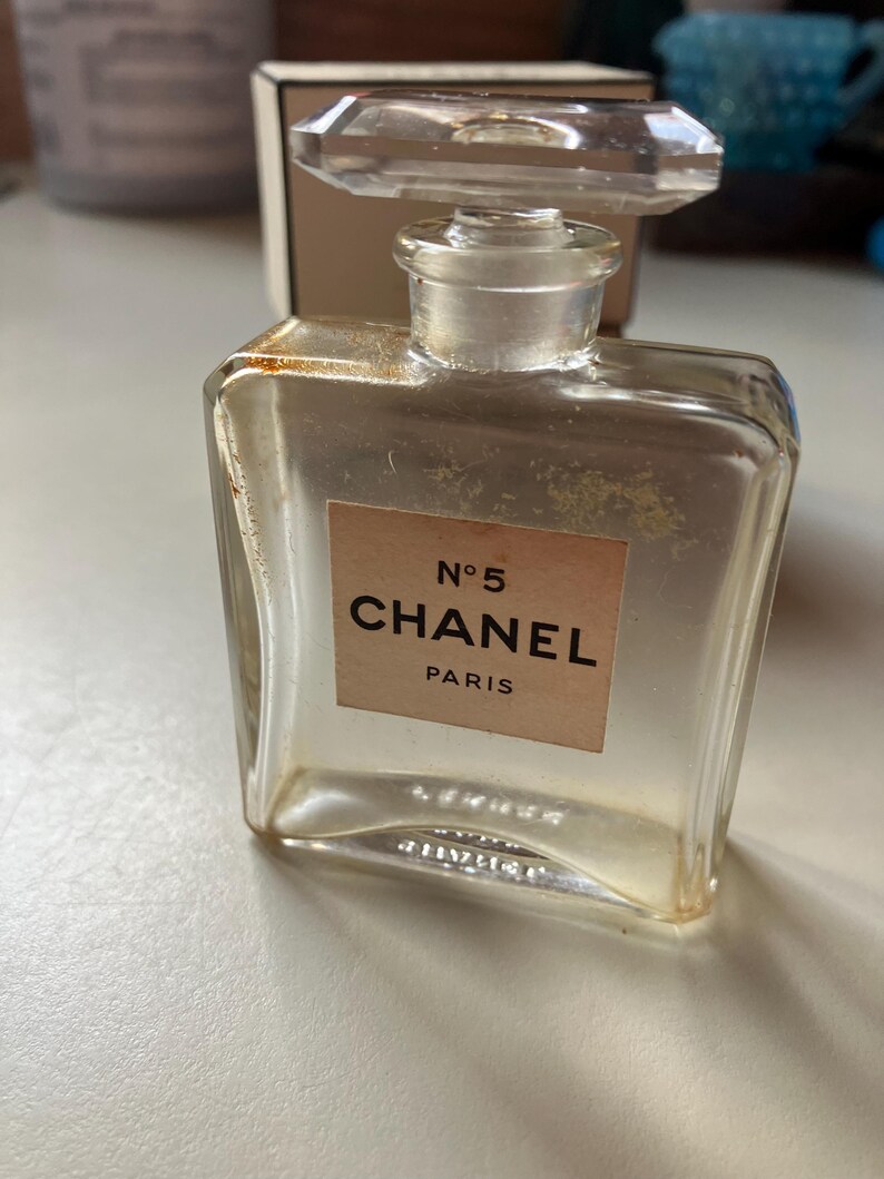 May include: A clear glass perfume bottle with a rectangular shape and a clear, faceted stopper. The label reads "N&deg;5 CHANEL PARIS" in black text on a beige background. The bottle appears to be vintage.