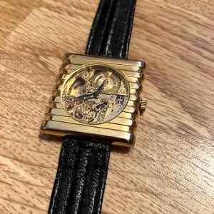 May include: A gold-toned rectangular wristwatch with a skeleton dial, showcasing the intricate inner workings. The watch has a black leather band. The watch is displayed on a wooden surface.