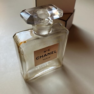 May include: A clear glass perfume bottle with a faceted stopper. The label reads "N&deg;5 CHANEL PARIS" in black text on a light brown background. A white and black box is in the background.