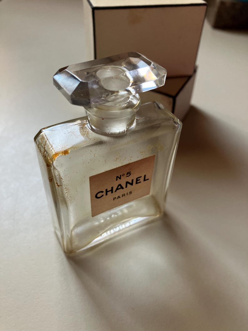 May include: A clear glass perfume bottle with a faceted stopper. The label reads "N&deg;5 CHANEL PARIS" in black text on a light brown background. A white and black box is in the background.
