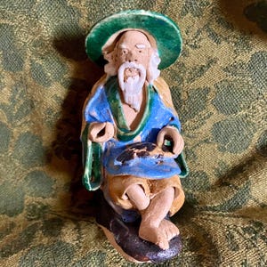 May include: A ceramic figurine of a seated man with a white beard and a green hat. He is wearing a blue robe with green accents and is holding a small object in his hands.
