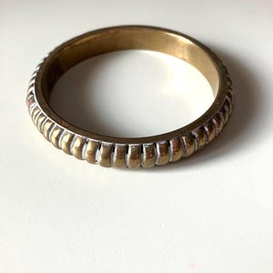 May include: A brass bangle bracelet with a ridged design. The bracelet has a gold finish with white accents on the ridged edges. The jewelry is circular and appears to be vintage.