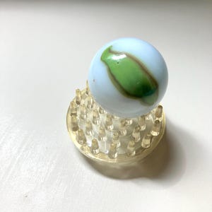 May include: A spherical glass marble with a milky white base and a prominent green and brown swirl pattern, resting securely on a clear circular plastic display stand featuring numerous small, upright, conical support pegs.