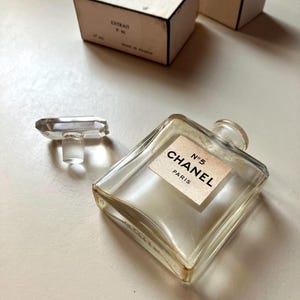 May include: A clear, square perfume bottle with a rectangular label that reads "CHANEL N&deg;5 PARIS." The bottle is next to a clear, rectangular stopper. Two small, rectangular boxes are in the background.