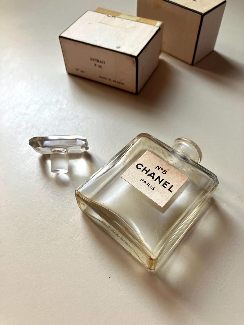 May include: A clear, square perfume bottle with a rectangular label that reads "CHANEL N&deg;5 PARIS." The bottle is next to a clear, rectangular stopper. Two small, rectangular boxes are in the background.
