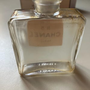 May include: A clear, vintage perfume bottle with a square shape and a rounded neck. The bottle has a label with the text "N 5 CHANEL PARIS" in a serif font. The bottle is empty and has a clear glass stopper.