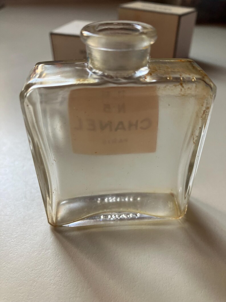 May include: A clear, vintage perfume bottle with a square shape and a rounded neck. The bottle has a label with the text "N 5 CHANEL PARIS" in a serif font. The bottle is empty and has a clear glass stopper.