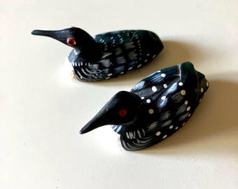 May include: Two hand-painted wooden loon figurines. The birds are black with white polka dots on their backs and white detailing on their chests. They have long black beaks and red eyes. The loons are approximately 4 inches long.