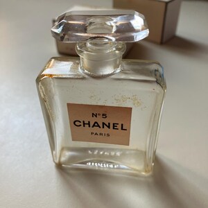May include: A vintage Chanel No. 5 perfume bottle. The clear glass bottle has a rectangular shape with a faceted, clear stopper. The label reads "N&deg;5 CHANEL PARIS" in black lettering on a beige background. The bottle is slightly aged.