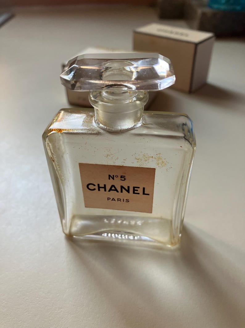 May include: A vintage Chanel No. 5 perfume bottle. The clear glass bottle has a rectangular shape with a faceted, clear stopper. The label reads "N&deg;5 CHANEL PARIS" in black lettering on a beige background. The bottle is slightly aged.