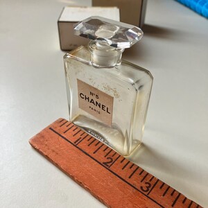 May include: A vintage Chanel No. 5 perfume bottle with a clear, rectangular design and a faceted, clear stopper. The label reads "N&deg;5 CHANEL PARIS". An orange wooden ruler is placed next to the bottle.