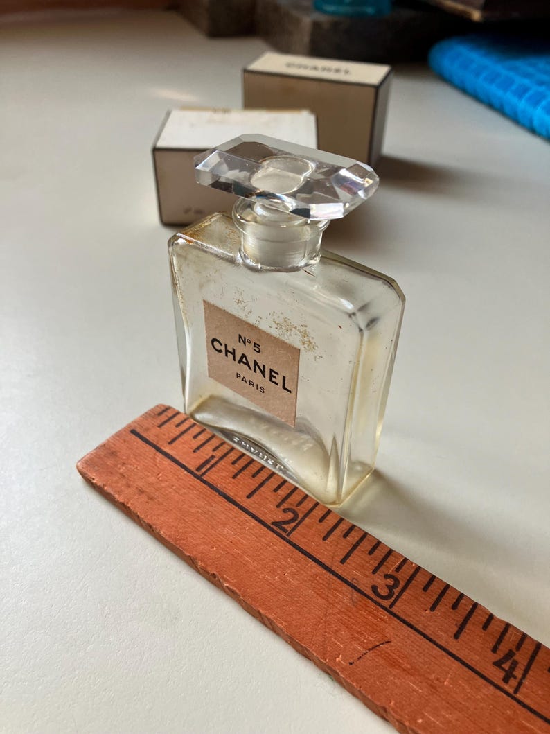 May include: A vintage Chanel No. 5 perfume bottle with a clear, rectangular design and a faceted, clear stopper. The label reads "N&deg;5 CHANEL PARIS". An orange wooden ruler is placed next to the bottle.