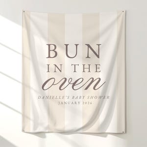 May include: A beige and white striped fabric banner with the words "BUN IN THE oven" in brown lettering. Below the text is "DANIELLE'S BABY SHOWER JANUARY 2026". The banner is hanging on a white wall.
