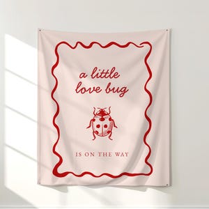 May include: A blush-colored wall tapestry with a red wavy border and text that reads "a little love bug is on the way." A red ladybug illustration is centered below the text. The words "blush background with red font" are at the bottom.