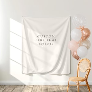 May include: Cream-colored custom birthday tapestry hanging on a white wall. The tapestry has the words "CUSTOM BIRTHDAY tapestry" printed on it. A bundle of balloons in white, pink, and rose gold colors is next to a wicker chair.