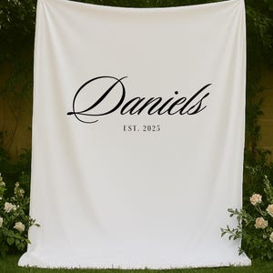 Può includere: Un fondale in tessuto bianco con il nome "Daniels" in elegante scrittura nera e il testo "EST. 2025" sottostante. Lo sfondo è appeso all'aperto, con vegetazione e fiori sullo sfondo.