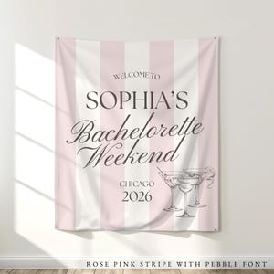 Pu&ograve; includere: Un banner a righe rosa e bianche con la scritta "WELCOME TO SOPHIA'S Bachelorette Weekend CHICAGO 2026". Il banner presenta anche un'illustrazione di due bicchieri da martini. Il banner &egrave; appeso a una parete bianca.