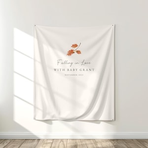 May include: A cream-colored fabric wall tapestry featuring a watercolor illustration of orange and brown autumn leaves at the top. The text reads Falling in Love WITH BABY GRANT NOVEMBER 2025 in a combination of script and serif fonts.