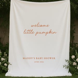 May include: A large white fabric backdrop featuring the orange text "welcome little pumpkin" in a cursive script, followed by the text "MADDY'S BABY SHOWER" and "JUNE 2026" in a clean, sans-serif font, displayed as a vertical banner.