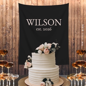 May include: A two-tiered white wedding cake with floral decorations sits on a gold-colored cake stand. Behind the cake is a black banner with the name "WILSON" and the year "est. 2026" in white. Champagne glasses are stacked on either side.