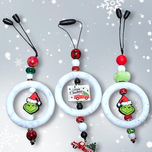 May include: Christmas-themed car charms. Each charm features a white marble-effect ring with a Grinch or a red truck with a Christmas tree. The charms have decorative beads and the text "Tis the season".