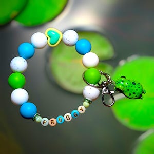 May include: A beaded bracelet with blue, white, and green beads, and a yellow heart-shaped bead. The bracelet has the word "Froggy" spelled out in letter beads. A green frog charm is attached to the bracelet with a silver clasp.