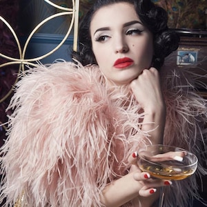 May include: A woman with dark hair and red lipstick wears a light pink feather boa and holds a champagne coupe. The background features a gold decorative element and a peacock feather pattern.