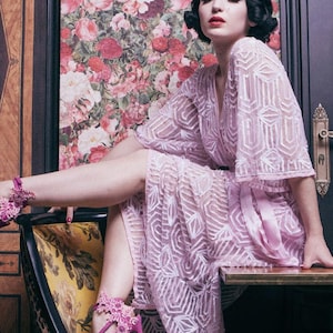 Puede incluir: Una bata lila estampada con cinturón a juego. La bata se combina con tacones altos rosas adornados con detalles florales. El fondo presenta papel pintado floral y un reloj vintage.