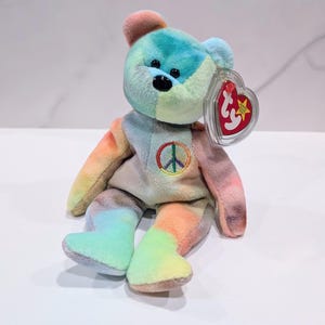 May include: A colorful plush bear with a peace sign on its chest. The bear features a rainbow tie-dye pattern on its fur, with a mix of blue, green, orange, and pink hues. It has black eyes and a red heart-shaped tag.