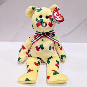 May include: A yellow plush bear with red, green, and white accents. The bear has a bow tie with red, white, and green stripes and a small tag. The bear is decorated with red and green shapes.