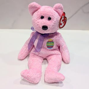 May include: A pink plush teddy bear with black eyes and a black nose. The bear has a purple ribbon around its neck and an embroidered Easter egg design on its chest. A red tag is attached to its ear.