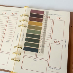 May include: A weekly planner with a brown leather cover and gold rings. The planner has a ruler with a colour guide for marking appointments. The ruler has a scale in centimetres. The planner has sections for Tuesday, Wednesday, Friday, Saturday, Sunday, and a weekly spread.