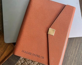 Puede incluir: Una agenda de cuero marrón con un cierre dorado y el texto "PONDER PAPER CO" en relieve en la parte delantera.