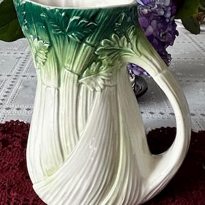 May include: A ceramic pitcher with a celery stalk design. The pitcher is primarily white with green accents on the top and handle. The handle is curved, and the pitcher has a wide opening. The pitcher is sitting on a burgundy crocheted surface.