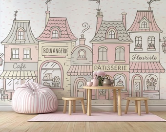 May include: A whimsical illustration of a French village scene with a cafe, bakery, and flower shop. The buildings are in soft pink, beige, and gray tones. A striped beanbag chair, small wooden table, and stools are in front of the mural. A pink rug is on the floor.