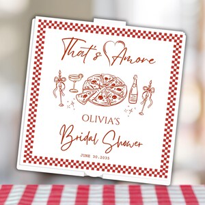 May include: A square white pizza box with a red checkered border. The lid features red line art of a pizza, a cocktail, a bottle, and two candles. Text reads: That's Amore, OLIVIA'S Bridal Shower, and the date JUNE 30, 2035, in red lettering.