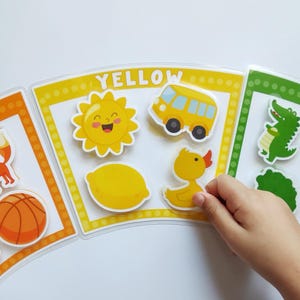 Puede incluir: Juego educativo de aprendizaje de colores con tarjetas naranja, amarilla y verde. Cada tarjeta muestra imágenes como un zorro, un sol, un autobús y un cocodrilo. La mano de un niño interactúa con la tarjeta amarilla, seleccionando una pegatina de pato amarillo.