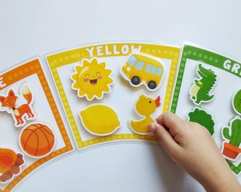 May include: Educational color learning set featuring orange, yellow, and green cards. Each card displays images like a fox, sun, bus, and crocodile. A child's hand interacts with the yellow card, selecting a yellow duck sticker.