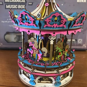May include: A wooden carousel music box with a pink, blue, and white color scheme. The carousel features three horses, a crown on top, and the words "MECHANICAL MUSIC BOX" on the box.