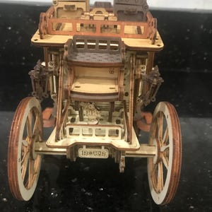 May include: A wooden model of a vintage carriage with large spoked wheels. The carriage is constructed from light and dark brown wood pieces, with intricate details. The word "ROKR" is visible on the back of the carriage.